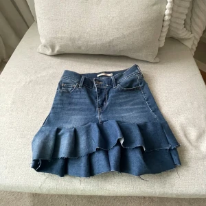 Levi's jeanskjol med volang - Supersnygg blå jeanskjol från Levi's med volangkant nertill och råa kanter. Klassisk femficksmodell med bälteshällor och dragkedja fram. Perfekt för dig som vill ha en cool och trendig denimlook.