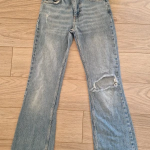 Ljusblå bootcut jeans från Gina Tricot - Säljer ett par ljusblå bootcut jeans från Gina Tricot, modell Perfect Jeans. De har en snygg sliten look med hål på ena knät och klassiska femficksdetaljer. Jeansen är i denim med låg midja och raka ben som går ut lite nertill.