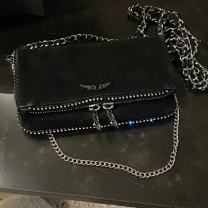 Zadig & Voltaire väska - Snygg svart axelväska i mocka från Zadig & Voltaire och snygga detaljer med stenar. Storlek ”nano”. Den är väl använd. Defekter förekommer som sminkfläckar (bild 5) men går att få bort säkert (därav priset), 3 stenar har lossnat men inget som syns alls. Dustbag medföljer inte eftersom hon hade tappat bort den när jag köpte här! 
