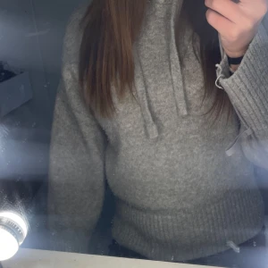 Trendig Grå stickad hoodie med snörning från Zara  - Mysig grå stickad hoodie med snörning i halsen och breda ribbade muddar. Perfekt för kyliga dagar och enkel att matcha med jeans eller leggings. Långärmad och i en klassisk loose passform som ger en avslappnad vibe.