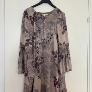 Odd Molly mönstrad långärmad klänning - Supersöt klänning från Odd Molly i beige med lila och grått blommönster. Långärmad modell med vida ärmar och knappar framtill. Skön och luftig passform, perfekt för dig som gillar boho-stil. Materialet känns mjukt och faller snyggt.