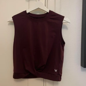 Vinröd tränings linne  - Säljer en vinröd ärmlös linnetopp från stadium med twistad detalj framtill. Toppen har rund halsringning och är tillverkad i mjukt, stretchigt material som passar perfekt till träning eller chill. Liten stadium-logga i vitt nedtill på framsidan.