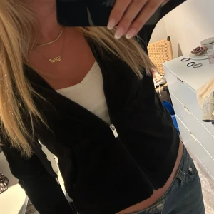 Svart velour hoodie med dragkedja - Svart hoodie i mjuk velour med dragkedja framtill och huva med snören. Jackan har två fickor fram och en croppad passform som sitter snyggt till jeans eller kjol. Perfekt för en chill och trendig look.