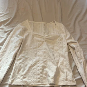 Vit blus med spets och v-ringning - Söt vit blus med lång ärm och v-ringad byst. Blusen har broderade detaljer och spetsmönster över hela tyget samt lätt rynkning vid bysten. Perfekt för dig som gillar romantisk och feminin stil.