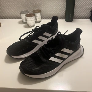 Svarta Adidas sneakers med vita ränder - Säljer ett par svarta Adidas sneakers med klassiska vita ränder på sidorna. Skorna har snörning, platt sula och är tillverkade i mesh och syntetmaterial. Perfekta för dig som gillar sportig stil och vill ha bekväma skor till vardagen.