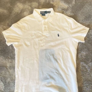 Vit pikétröja från Polo Ralph Lauren - Klassisk vit pikétröja från Polo Ralph Lauren med korta ärmar och den ikoniska broderade loggan på bröstet. Tröjan har krage och knäppning med två knappar. Tillverkad i mjuk bomull som känns skön mot huden. Perfekt för en clean och stilren look.