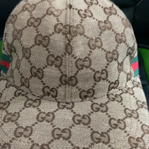 keps med GG-mönster - Snygg keps  i beige och brunt med klassiskt GG-monogram över hela kepsen. Sidorna har gröna och röda ränder som ger den ikoniska Gg-looken. Tillverkad i bomullsmix med vit insida och märkning på etiketten.