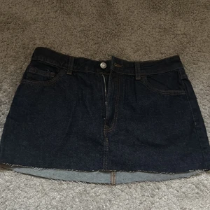 Mörkblå jeanskjol med rå kant - Snygg mörkblå jeanskjol med klassiska fickor och rå, fransig kant nertill. Kjolen har dragkedja och knapp framtill samt bälteshällor. Perfekt för en chill och trendig look.