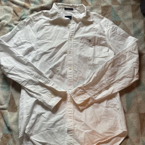 Vit oxfordskjorta från Gant - Klassisk vit oxfordskjorta från Gant i bomull. Skjortan har lång ärm, bröstficka med broderad Gant-logga och knappar framtill. Regular fit med snygg krage, perfekt till jeans eller chinos. Enkel och stilren design som alltid funkar. Skulle säga att den är lite stor i storlek