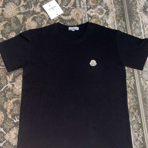 Svart Moncler t-shirt med logga - Svart t-shirt från Moncler med klassisk rund hals och liten broderad logga på bröstet. Enkel och stilren design som passar perfekt till jeans eller shorts. Tillverkad i mjuk bomull för skön känsla hela dagen.