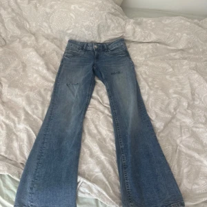 Blå bootcut jeans med slitningar - Säljer ett par blå bootcut jeans med lätt slitna detaljer vid knäna. Jeansen har klassisk femficksdesign, normal midja och är tillverkade i mjukt denimtyg. Perfekta för dig som gillar en retro vibe och vill ha ett par jeans med lite extra vidd nertill.