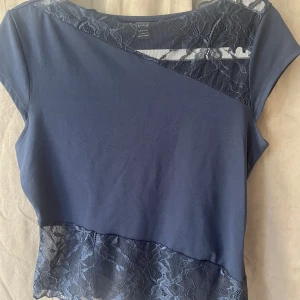 Mörkblå topp med spets från SHEIN - Snygg mörkblå topp från SHEIN med korta ärmar och dekorativa spetsdetaljer upptill och nertill. Toppen har en asymmetrisk design med blommig spets som ger en cool och feminin vibe. Perfekt att styla med jeans eller kjol för en trendig look.