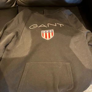 Svart hoodie från Gant med logga - Svart hoodie från Gant med broderad logga och sköld med stjärnor och ränder på bröstet. Klassisk känguruficka framtill och mjuk insida. Perfekt för dig som gillar stilren streetwear med sportig känsla.