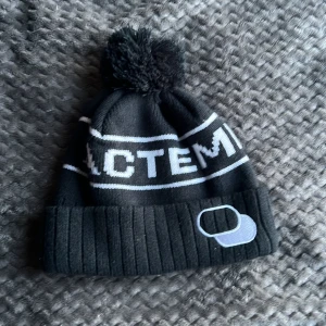 Svart mössa med tofs och vit text - Svart ribbstickad mössa med stor tofs på toppen och vit text runt om. Mössan har en bred uppvikt kant och en vit broderad detalj framtill. Perfekt för kalla dagar och snygg till streetwear.