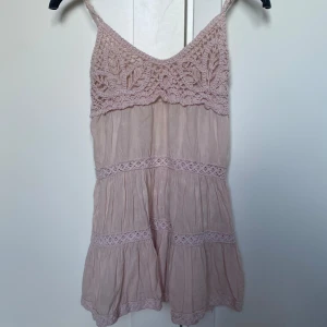 Ljusrosa spetslinnetopp JS Millenium - Supersöt ljusrosa linnetopp från JS Millenium med virkade spetsdetaljer upptill och längs med volangerna. Tunt, luftigt tyg och smala axelband. Perfekt för varma dagar när du vill ha en romantisk och bohemisk vibe.