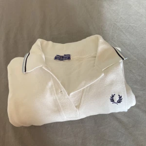 Vit pikétröja från Fred Perry - Klassisk vit pikétröja från Fred Perry med blå broderad logga på bröstet och stilren krage med mörkblå rand. Tröjan är i bomull och har en tidlös design som passar perfekt till jeans eller shorts. Storlek: cirka 160, skick: 9/10, kontakta vid frågor!