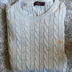 Ljusgrå kabelstickad tröja Morris XL - Kabelstickad tröja från Morris i ljusgrått, tillverkad i mjuk bomull. Klassisk rund halsringning och snyggt broderad logga i orange på bröstet. Perfekt för dig som gillar stilren och tidlös design.                       (Passform som en L) hör av dig vid frågor! 