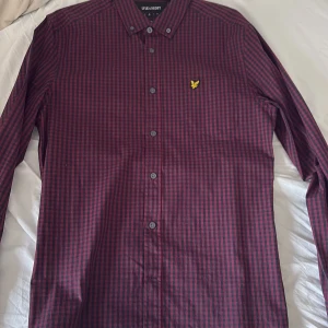Rutig vinröd skjorta från Lyle & Scott - Snygg vinröd och svart rutig skjorta från Lyle & Scott i slim fit-modell. Skjortan har långa ärmar, klassisk krage och knäppning framtill. Ikonisk gul logga broderad på bröstet. Perfekt för dig som gillar stilrena detaljer och en smalare passform. Passar dig som bär storlek M 