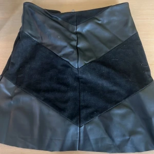 Svart kjol i skinn och mocka - Snygg svart kortkjol med coola paneler i både skinn och mocka. Kjolen har en stilren design med geometriska former och passar perfekt till en trendig outfit. Enkel att matcha med både boots och sneakers.