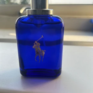 Ralph Lauren Polo Blue parfym - Snygg parfymflaska från Ralph Lauren i djupblått glas med silverdetaljer och den klassiska polospelaren i silver på framsidan. Flaskan har en stilren, fyrkantig form och metallisk spraykork. Perfekt för dig som gillar exklusiva dofter och ikonisk design.