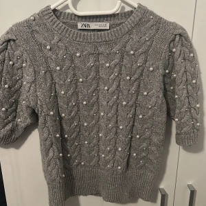 Grå stickad tröja med pärlor från Zara - Superfin grå stickad tröja från Zara med kortare ärmar och snyggt flätmönster. Tröjan är dekorerad med pärlor över hela och har ribbade muddar. Perfekt för dig som vill ha något extra till hösten eller vintern! 💕