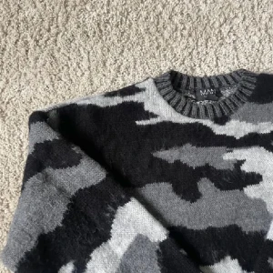 Grå camo stickad tröja från MAN - Snygg stickad tröja från MAN med grått och svart camouflage-mönster. Tröjan har rund halsringning, ribbade muddar och är långärmad. Perfekt för dig som gillar streetstyle och vill sticka ut lite extra.