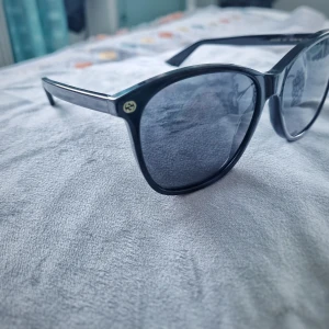 Svarta solglasögon från Gucci - Snygga svarta solglasögon från Gucci med klassisk, lite rundad fyrkantig form. Tjocka bågar i blank plast och diskret Gucci-logo i guld på sidan. Perfekt accessoar för att lyfta din stil.