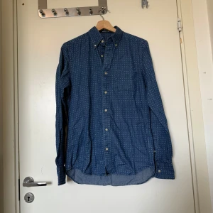 Blå mönstrad skjorta från GANT - Snygg blå skjorta från GANT i modellen Manhattan Indigo. Skjortan har små vita mönster, klassisk krage och knäppning framtill. Tillverkad i mjuk bomull och har långa ärmar med knappar vid manschetterna. Perfekt för dig som gillar stilrena detaljer. Storlek S. Mycket fint skick, jag hittar inga synlig anmärkningar eller defekter.