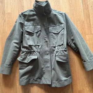 Grön parkajacka med stora fickor - Snygg grön parkajacka med hög krage och fyra stora fickor framtill. Jackan har dragsko i midjan och stängs med dold dragkedja. Mycket sparsamt använd. Upplevs som större än en 32:a. 