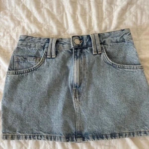 Ljusblå minikjol - Snygg ljusblå jeanskjol i kort modell med klassiska fickor fram och bak. Kjolen har hällor och matchande jeansbälte med metallspänne. Perfekt för en chill och trendig look.