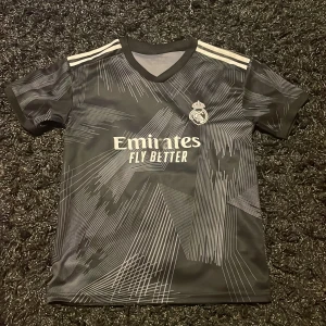Real Madrid Modric svart bortatröja - Snygg svart Real Madrid bortatröja med Modric 10 på ryggen. Tröjan har korta ärmar, V-ringning och vita Adidas-ränder på axlarna. Mönstret är grafiskt med tunna vita linjer. Materialet är lätt och andas, perfekt för fotboll eller träning.