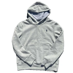 Grå hoodie från Polo Ralph Lauren - Snygg ljusgrå hoodie från Polo Ralph Lauren med klassisk broderad logga på bröstet. Tröjan har huva, känguruficka och är tillverkad i mjuk bomullsmix. Perfekt för chill dagar och enkel att matcha med jeans eller joggers.