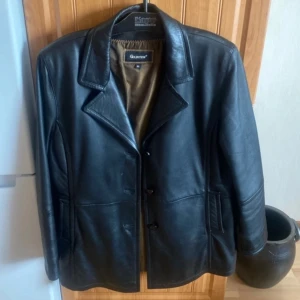 Äkta Svart skinnjacka från Goldstein - Stilren svart äkta skinnjacka från Goldstein med klassisk krage och knäppning framtill. Jackan har raka linjer, två fickor och är helfodrad med brunt innerfoder.