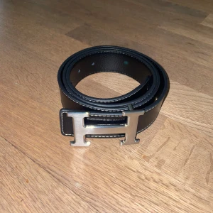 Svart H bälte - Ett svart bälte med H loggan på. Bältet är 115 cm långt, man får med en hålpipa som gör att man kan göra egna hål. Perfekt accessoar för att lyfta vilken outfit som helst.