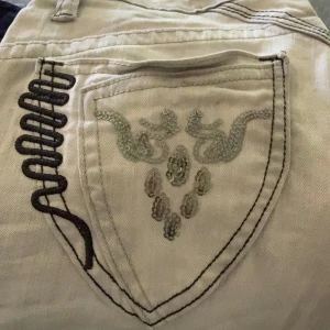 Ljusgrå jeans Miss Capri strl 26 - Snygga ljusgrå jeans från Miss Capri med broderade detaljer och paljetter på bakfickan. Byxorna har låg midja och raka ben, Perfekta för dig som gillar unika detaljer och en cool look.