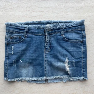 Blå jeanskjol med fransad kant - Snygg blå jeanskjol med slitna detaljer och fransad kant både upptill och nertill. Klassisk femficksmodell med bälteshällor och råa kanter för en cool, avslappnad vibe. Perfekt för dig som gillar streetstyle och denim.