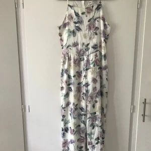 Lång blommig jumpsuit från H&M - Superfin lång jumpsuit från H&M med blommigt mönster i lila, grönt och beige på vit botten. Klänningen har smala axelband och är ärmlös, perfekt för sommarens varma dagar. Luftigt material som känns skönt mot huden.