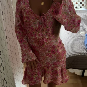 Blommig rosa kortklänning från Zara - Supersöt kortklänning från Zara med blommigt mönster i rosa och vita toner. Klänningen har långa ärmar, volang nertill och en snygg snörning vid urringningen. Perfekt för sommarens alla tillfällen!