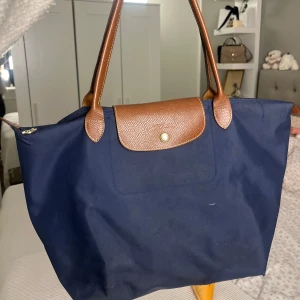 Marinblå Longchamp axelväska - Säljer en marinblå longchamp väska som är stor och perfekt att få plats mycket saker i. Älskar den här väskan passar verkligen till allt och så rymlig! Som man ser på bilden är väskan lite sliten vid botten hörnen därav det låga priset💗💗