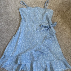 Blå omlottklänning med volang - Söt ljusblå omlottklänning med vitt blommönster och volangkant. Klänningen har smala axelband, knyt i midjan och en feminin passform. Perfekt för varma dagar och somriga vibes. Storlek 140🥰
