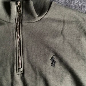 Olivgrön half zip tröja från Polo Ralph Lauren - Snygg olivgrön långärmad tröja från Polo Ralph Lauren med half zip och broderad logga på bröstet. Tröjan har en hög krage och är gjord i mjuk bomull, perfekt för lager-på-lager-stil. Enkel och stilren design som funkar året runt.