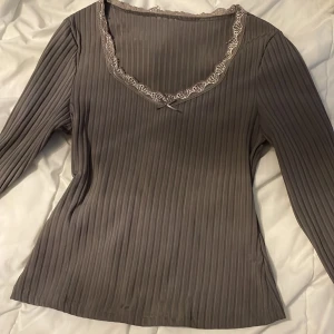 Ribbad grå långärmad topp med spets - Supersnygg ribbad grå långärmad topp med v-ringning och spetsdetaljer vid halsringning och ärmslut. Smal passform och diskret liten rosett framtill. Perfekt för dig som gillar stilrena och feminina detaljer.