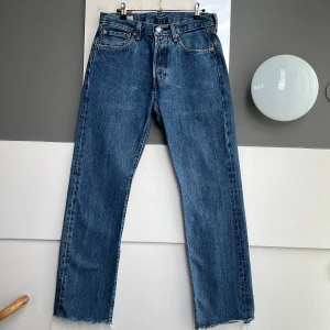 Levi's 501 raka jeans med rå kant - Klassiska blå Levi's 501 jeans med rak passform och hög midja. Jeansen har fem fickor, knappgylf och rå, avklippt fåll nedtill för en cool, avslappnad look. Tillverkade i robust bomullsdenim med ikonisk röd Levi's-tag på bakfickan. L34 original, men nerklippt till L31.
