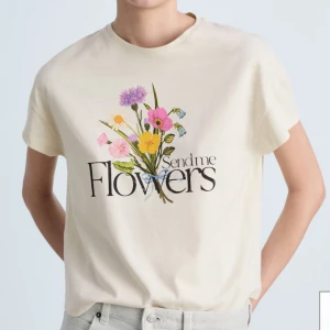 Blommig t-shirt med tryck  - Krämvit t-shirt med färgglatt blommotiv och texten 'Send me Flowers' på bröstet. T-shirten har klassisk passform, rund hals och korta ärmar. Materialet är mjuk bomull som känns skönt mot huden. Perfekt för dig som gillar en söt och lekfull stil. Finns en fläck, därav priset 