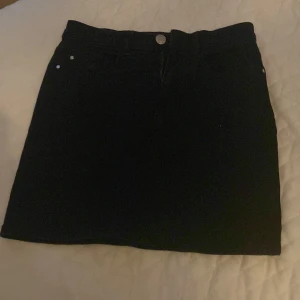 Svart jeanskjol med klassisk look - Svart kort jeanskjol med klassisk femficksmodell och knapp framtill. Snygg och enkel design som passar till det mesta. Rak passform och midjehög. Perfekt för dig som gillar en stilren och tidlös vibe.