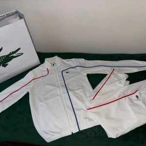Vita Lacoste tracksuit med röd och blå detalj - Säljer ett tracksuit från Lacoste med stilrena blå och röda ränder längs sidorna och den klassiska krokodilloggan på benet. Byxorna har resår i midjan och vid bensluten för optimal passform och komfort – perfekta för dig som gillar en sportig men clean stil.  Storlek: S, men passar även M tack vare den stretchiga midjan. Byxorna är helt oanvända och har endast legat i garderoben. Nypris: 2700 kr Säljes för: 2100 kr – priset kan diskuteras vid snabb affär. Kvitto finns. 