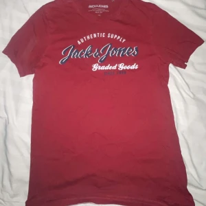 Röd t-shirt Jack & Jones M - Röd t-shirt från Jack & Jones med tryckt text i vitt och blått på bröstet. Klassisk rund halsringning och korta ärmar. Skön och mjuk bomullskvalitet, perfekt till jeans eller shorts.