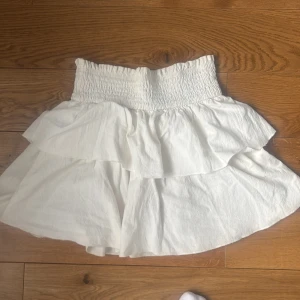 Vit volangkjol i bomull - Supersöt vit kortkjol i 100% bomull med bred smock i midjan och dubbla volanger. Perfekt för dig som gillar en luftig och flowy stil. Kjolen är enkel att matcha och har en riktigt fräsch vibe.