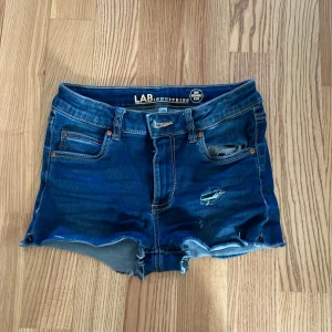 Blå jeansshorts från LAB Industries - Snygga blå jeansshorts från LAB Industries med slitna detaljer och råa kanter. Klassisk femficksmodell med dragkedja och knapp. Perfekta för sommaren och ger en cool, avslappnad vibe. Tillverkade i hållbar denim.