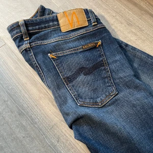 Nudie jeans - Snygga mörkblå  jeans från Nudie med klassisk femficksdesign och orangea sömmar. Jeansen har en diskret tvättad look och Nudie-logga på bakfickan samt läderpatch i midjan. Perfekta för dig som gillar en skön passform och stilren look.